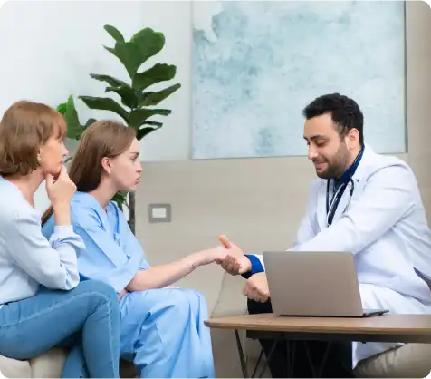A doctor in a white coat shaking hands with a patient in a hospital gown, while another person sits nearby. A laptop is open on the table, and a plant is visible in the background.
