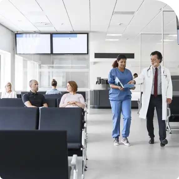 A doctor speaks with a nurse in a waiting room, surrounded by chairs and medical posters, conveying a sense of care.