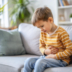 Child sitting on a sofa, holding their stomach with a pained expression, possibly experiencing abdominal discomfort or stomach pain at home.