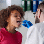 Doctor examining a child’s throat using a tongue depressor during a check-up, possibly performing a strep throat test in a clinical setting.