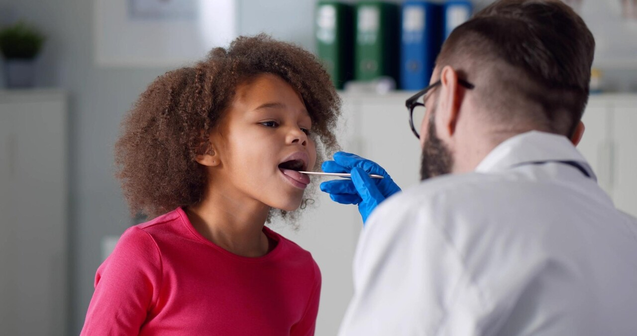 Doctor examining a child’s throat using a tongue depressor during a check-up, possibly performing a strep throat test in a clinical setting.