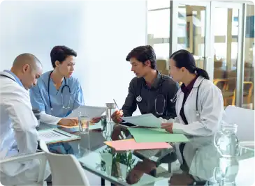 A group of doctors engaged in discussion around a conference table, with medical charts and laptops in front of them.