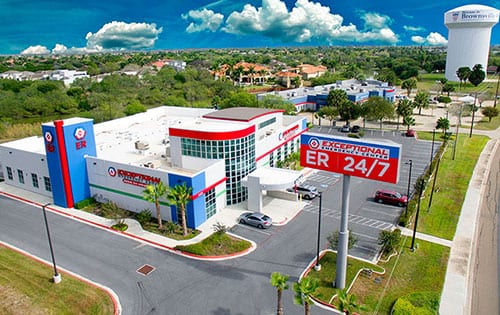An aerial view of the "Exceptional Emergency Center" featuring a large "ER 24/7" sign. The building has a modern design with blue and red accents, surrounded by a parking lot and greenery. A water tower is visible in the background, along with residential areas.