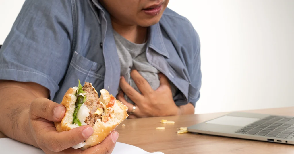 A man sitting at a desk with a sandwich in hand and a laptop open, showing signs of chest pain after eating.