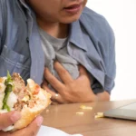 A man sitting at a desk with a sandwich in hand and a laptop open, showing signs of chest pain after eating.