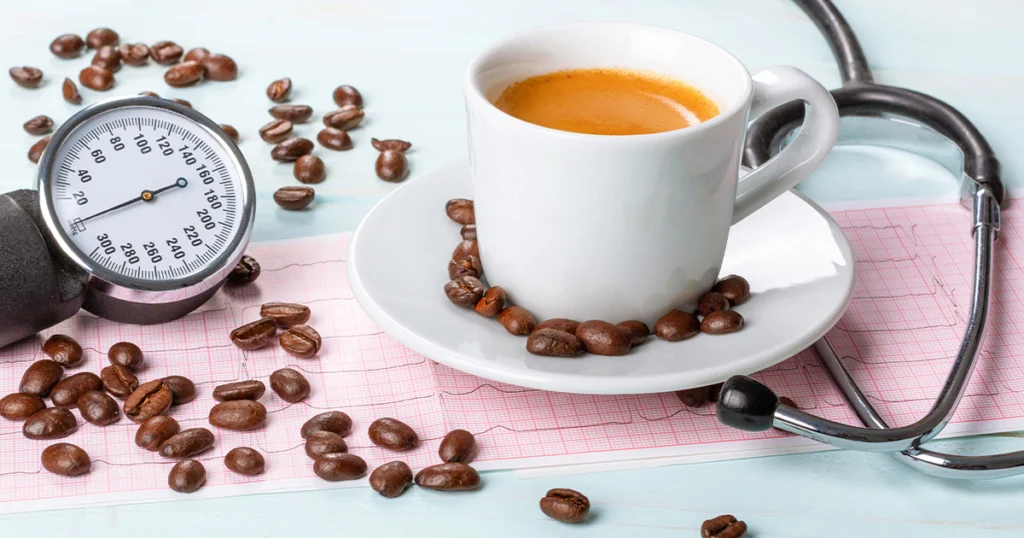 Stethoscope beside a cup of coffee, coffee beans, and a blood pressure monitor, representing the link between coffee and high blood pressure.