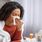 A sick woman wrapped in a blanket uses a tissue while medicine bottles sit nearby, representing symptoms of Flu A vs Flu B