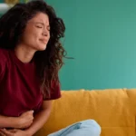 A woman sitting on a couch holding her stomach in pain.