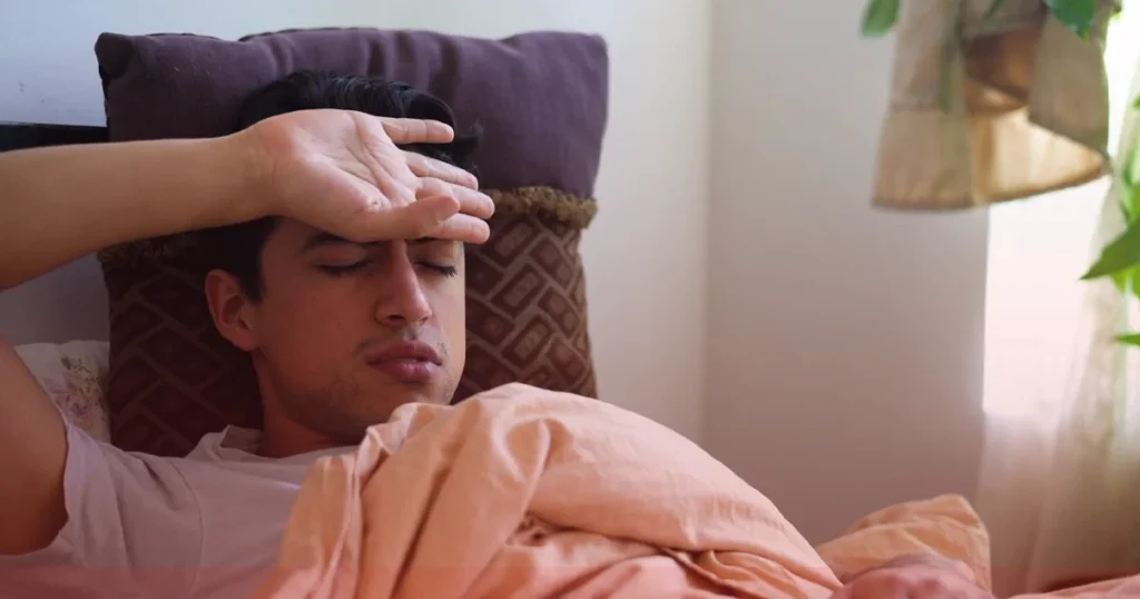 Man lying in bed with his hand on his forehead, appearing unwell with fever symptoms while resting under a blanket at home.