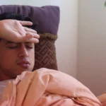 Man lying in bed with his hand on his forehead, appearing unwell with fever symptoms while resting under a blanket at home.