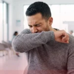 A realistic stock photo of a man in a modern living room, coughing into the crook of his elbow with a dry, irritated expression, representing a persistent dry cough.