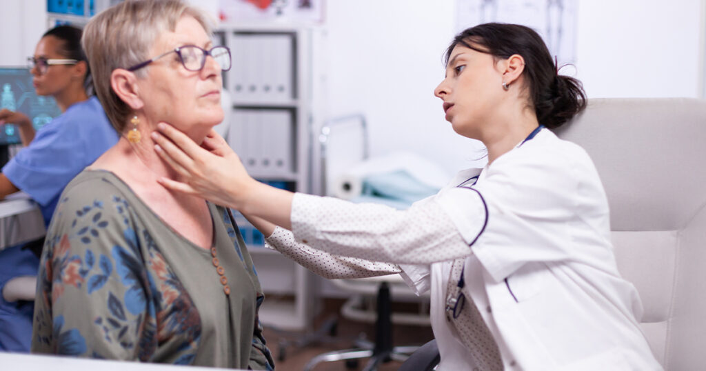 A doctor in a white coat examines the neck of an older woman with glasses during a medical consultation.