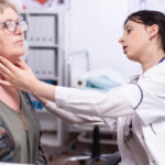 A doctor in a white coat examines the neck of an older woman with glasses during a medical consultation.