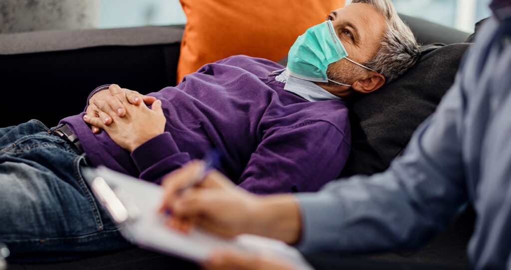 A man wearing a face mask lies on a couch while someone takes notes in the foreground.