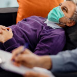 A man wearing a face mask lies on a couch while someone takes notes in the foreground.