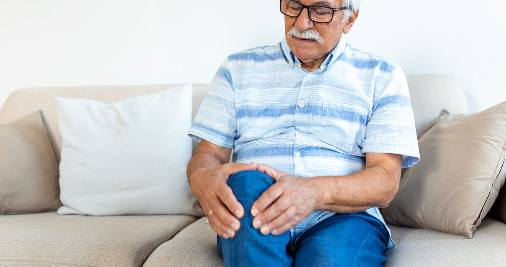 Senior man holding his painful knee while sitting on sofa.