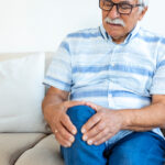 Senior man holding his painful knee while sitting on sofa.