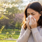 Woman sneezing into a tissue near blooming trees during pollen season