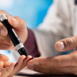 A healthcare professional assists a patient with a blood glucose finger-prick test using a lancing device.