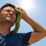 Man wiping sweat with a towel under bright sunlight on a hot day.