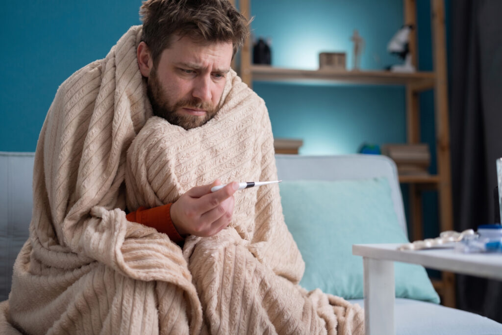 A sick man wrapped tightly in a thick beige blanket sitting on a couch and checking his temperature with a digital thermometer.