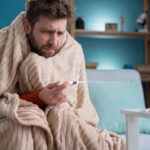 A sick man wrapped tightly in a thick beige blanket sitting on a couch and checking his temperature with a digital thermometer.