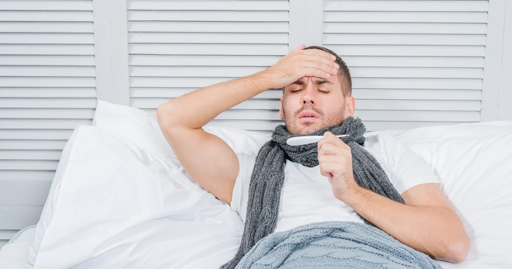 A sick man in bed is checking his temperature with a thermometer.