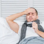 A sick man in bed is checking his temperature with a thermometer.