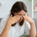 Woman sitting on a grey couch covering her eyes in discomfort due to dry eye irritation.