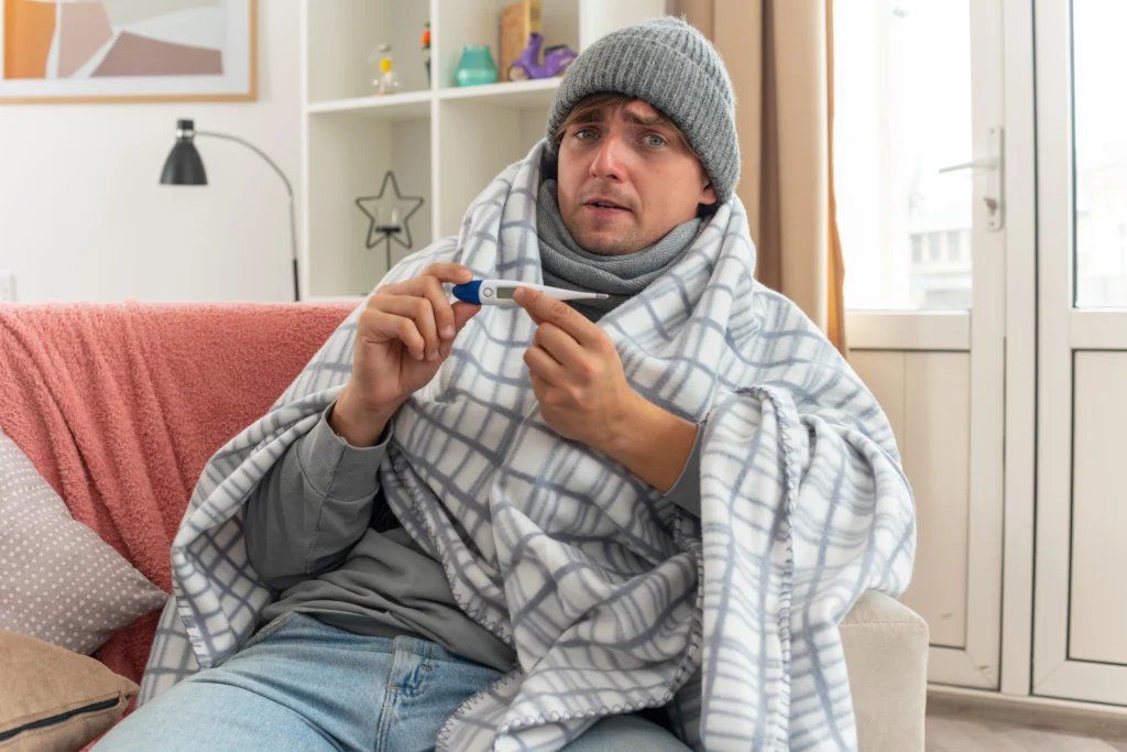 Sick man wrapped in a blanket and wearing a beanie, holding out a digital thermometer.