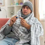 Sick man wrapped in a blanket and wearing a beanie, holding out a digital thermometer.