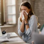 A woman blowing her nose into a tissue at her work desk.