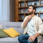 Man sitting on a sofa holding his neck in pain from a sore throat.
